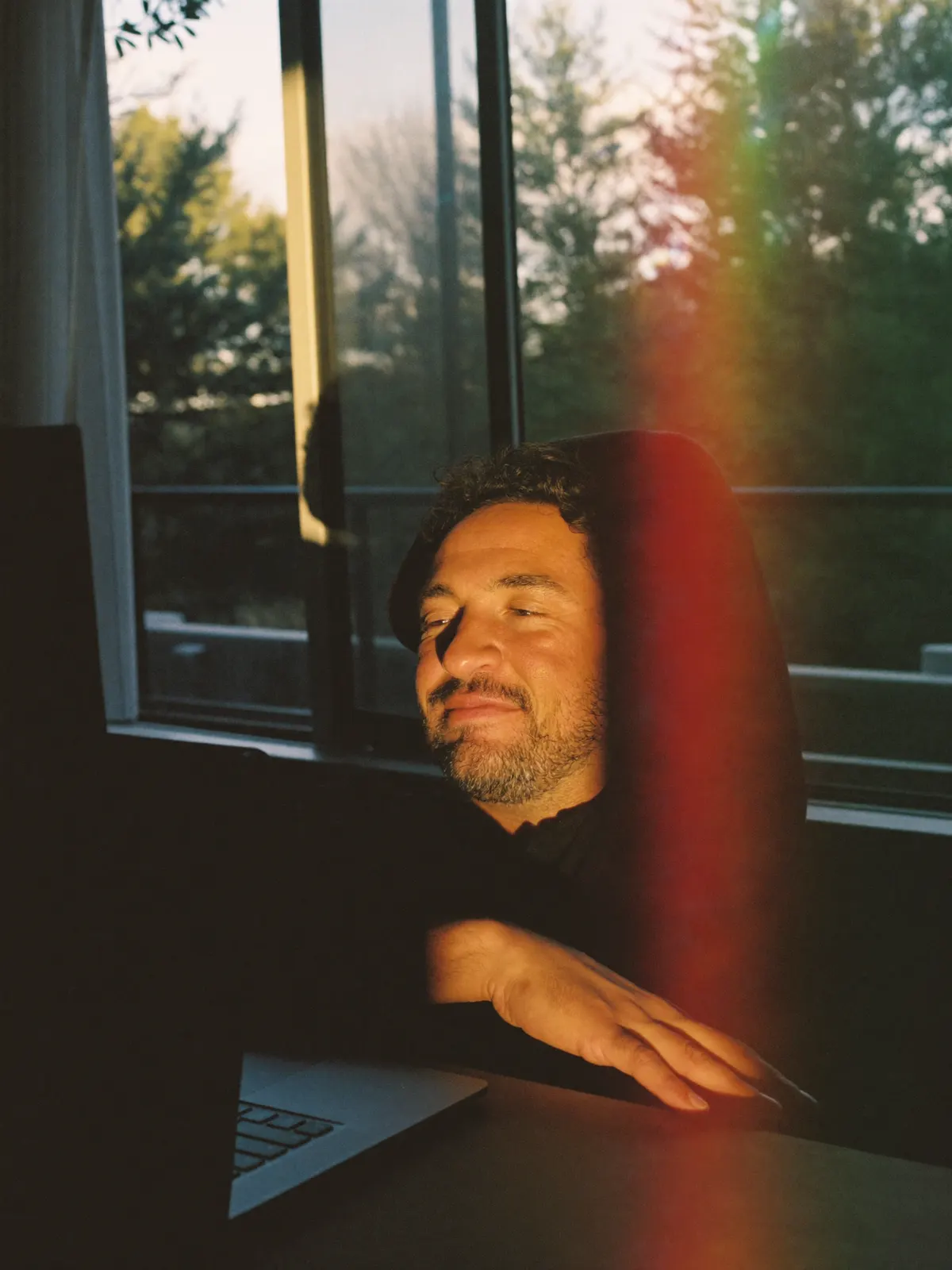 A man at his desk at the end of the day, eyes closed and smiling, a band of prism light beside him
